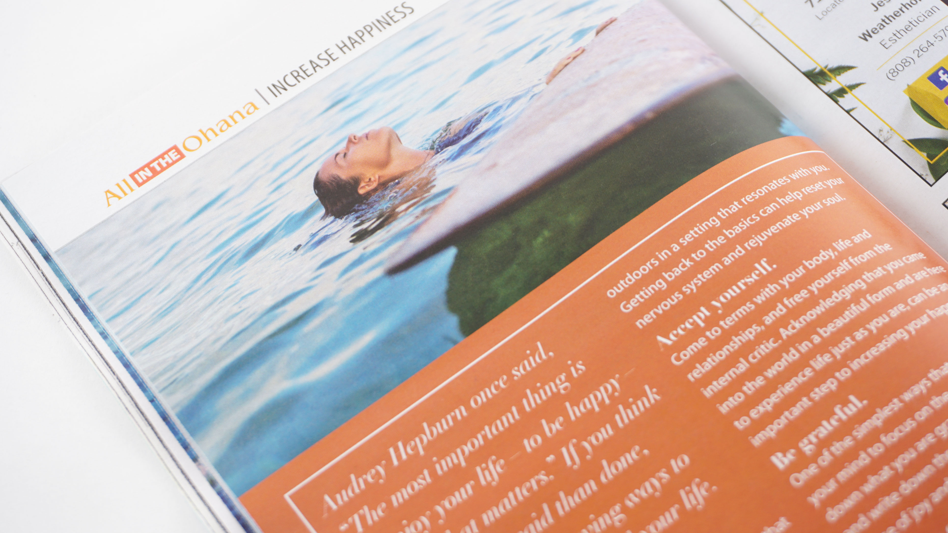 Picture of a magazine article with an image of a woman leaning her head back in the ocean, with her hands resting on a surfboard in front of her. Beneath the image is a box reddish brown box with white text from the Maui Family Magazine article, "Simple Ways to Increase Happiness Every Day," designed by Alexa Lasco.